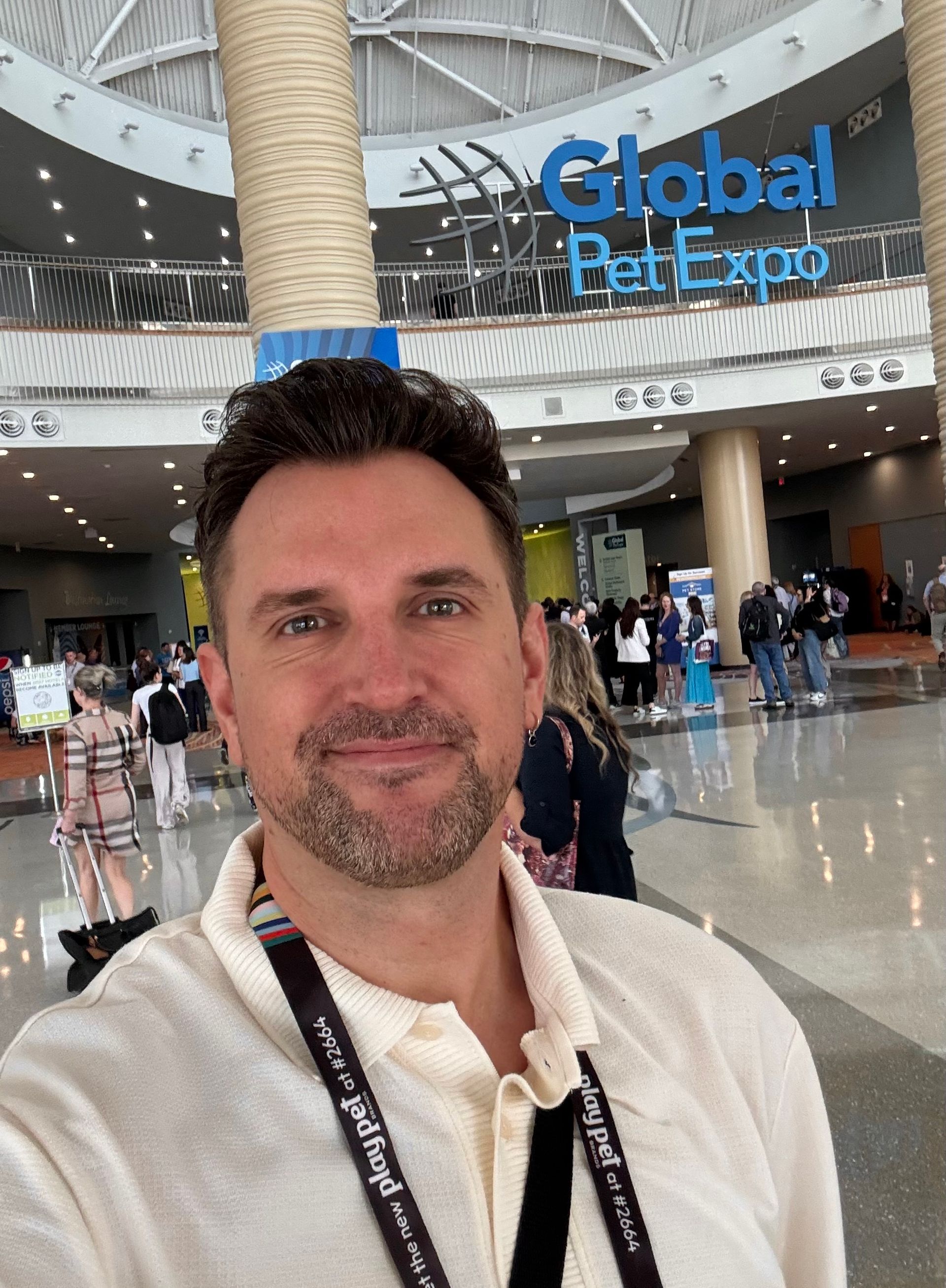 A smiling person kneels to shake hands with a golden dog wearing a yellow bandana, with a second dog nearby indoors.