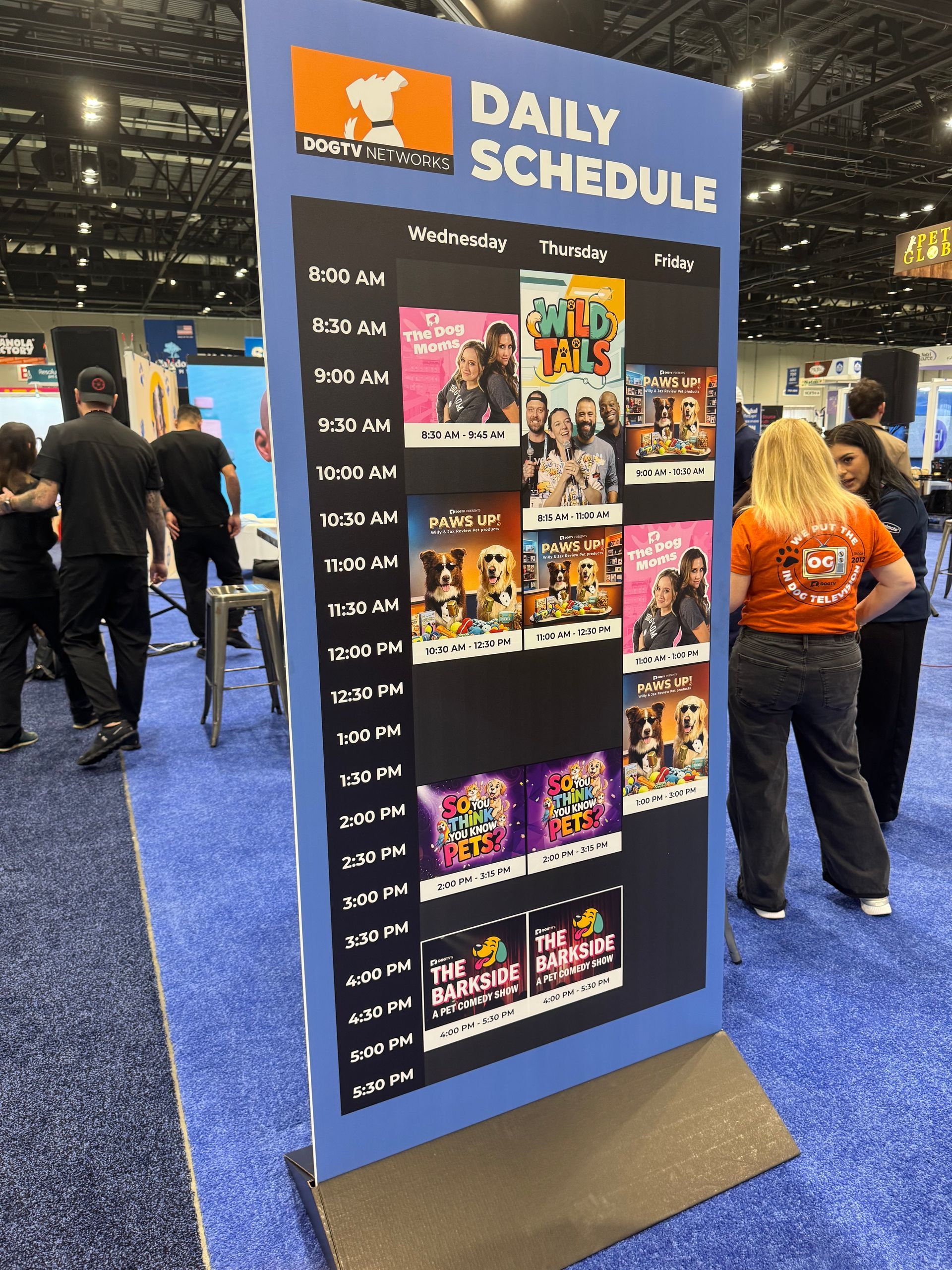 A smiling man and woman pose for a selfie at a crowded indoor event with green carpeting and decorated stalls.