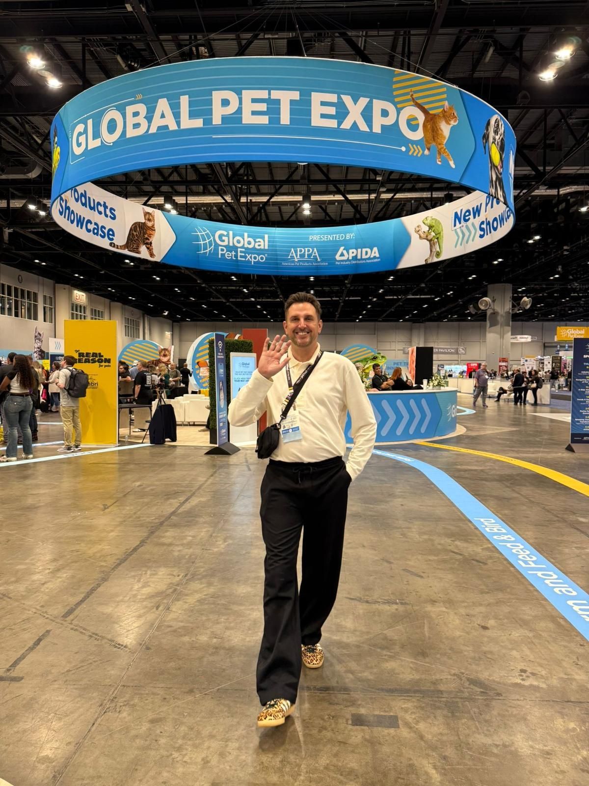 A smiling person kneels to shake hands with a golden dog wearing a yellow bandana, with a second dog nearby indoors.