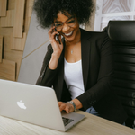 A professional smiling while talking on a smartphone and typing on a laptop at a desk in a modern office.