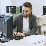 A person in a gray blazer smiling while typing on a computer in an office.