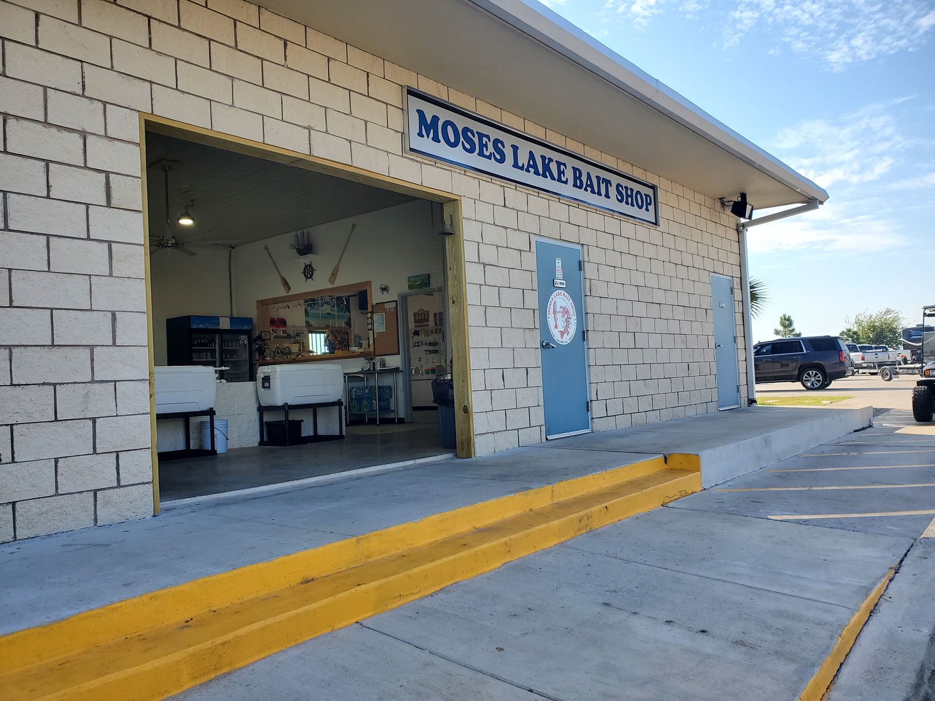 Concrete boat ramp leading to a waterway. A building with a sign and blue door on the right. Blue sky and clouds overhead.