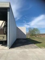 RV parked under a metal shelter beside a light brick wall, against a blue sky.