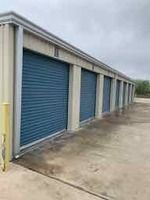 Storage units with blue doors in a row. Concrete ground, beige building, overcast sky.