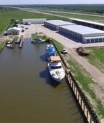 Boats docked in a canal near storage buildings and a highway in a green field.