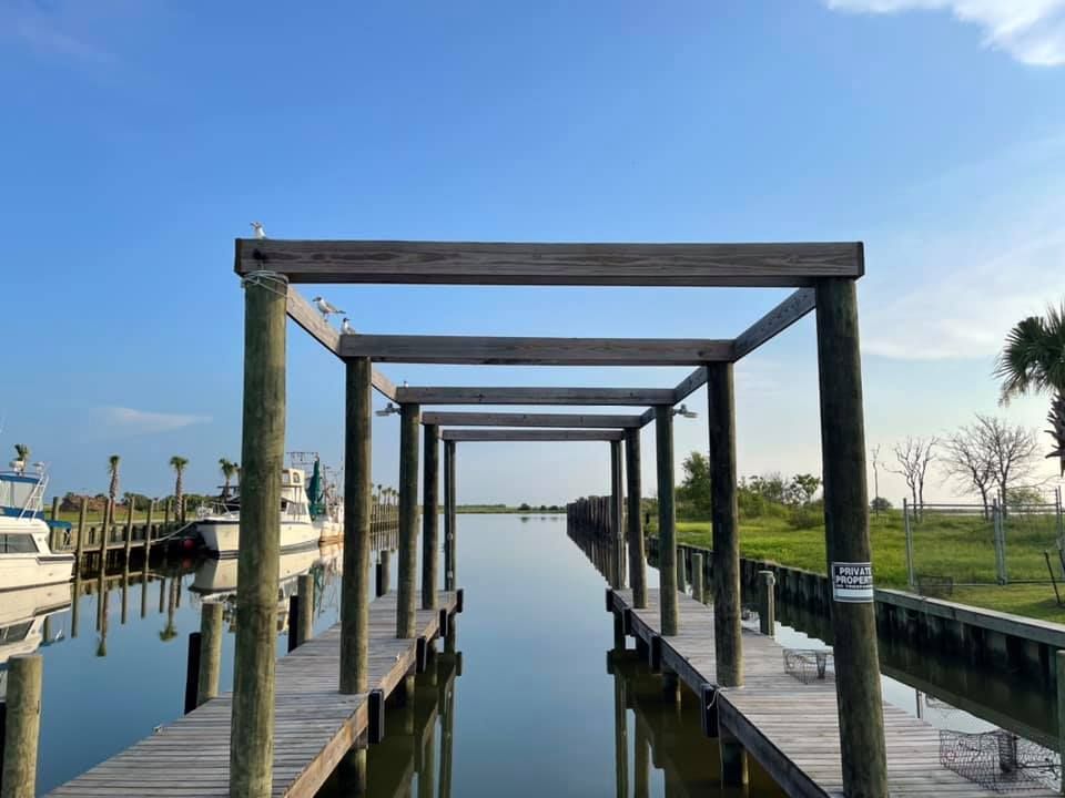Wooden dock with open-air rectangular frames extending over calm water channel, boats on left, green foliage on sides.