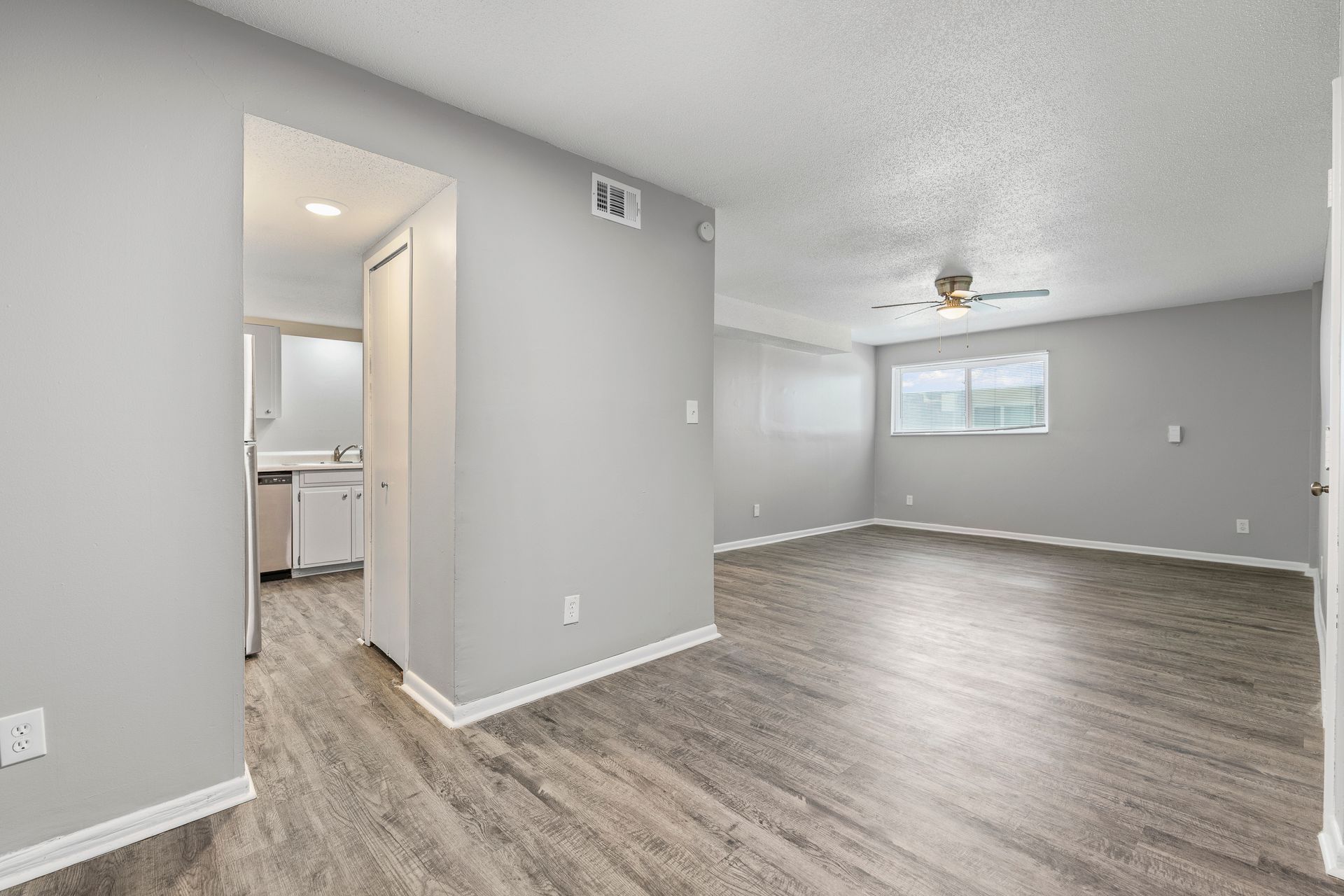 Interior view of a gray-walled apartment with wood-look flooring, a kitchen in the background, and a window on the right.