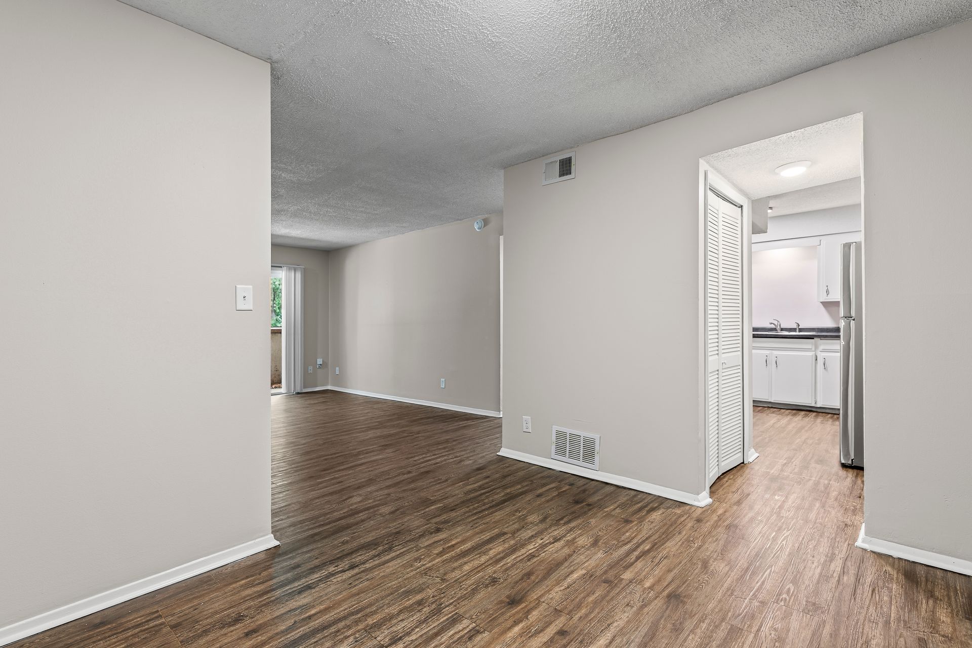 Empty apartment interior with wood-look floors, light gray walls, and an open doorway to a kitchen.
