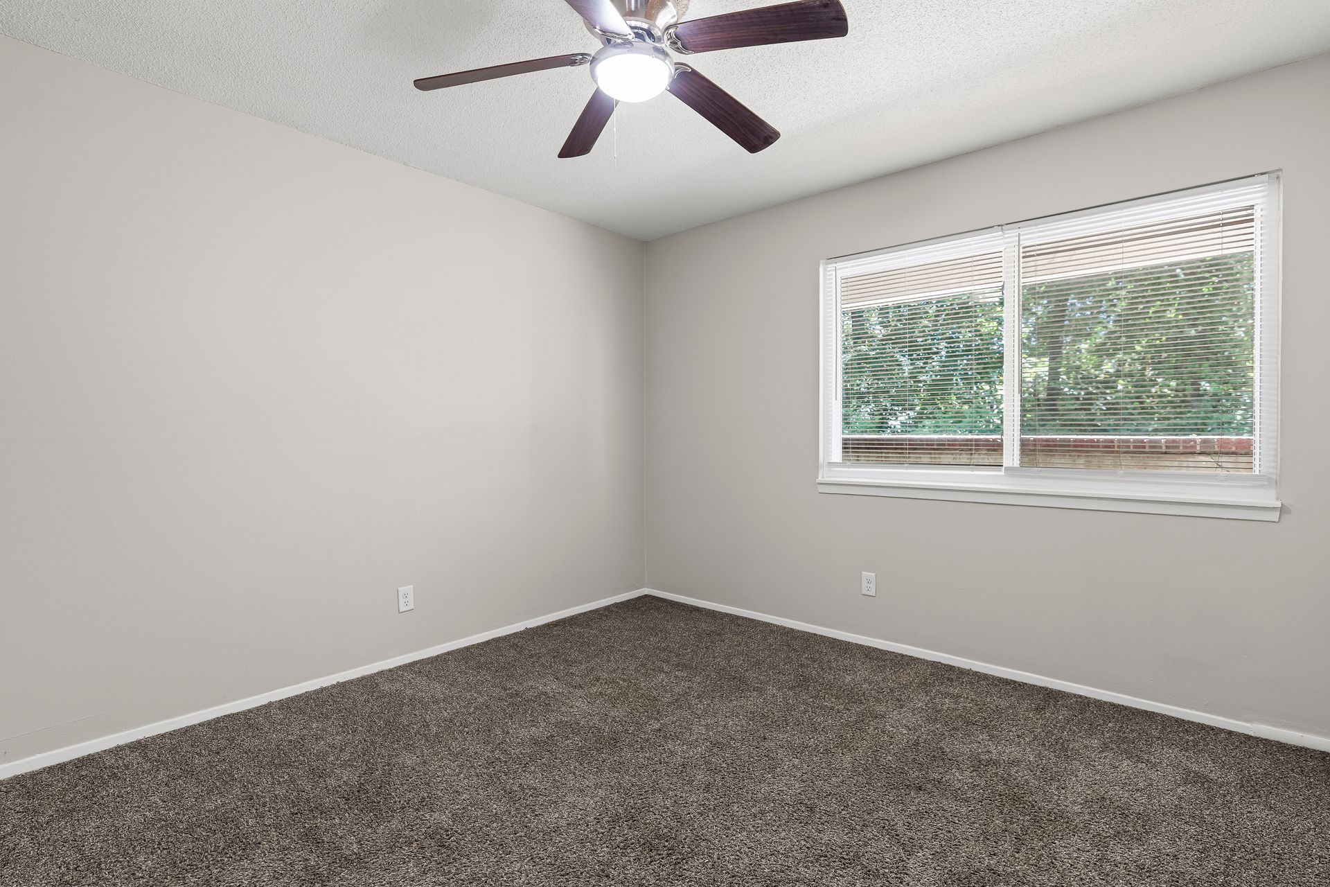 Empty bedroom with gray carpet, walls, and a ceiling fan. Window with greenery view.