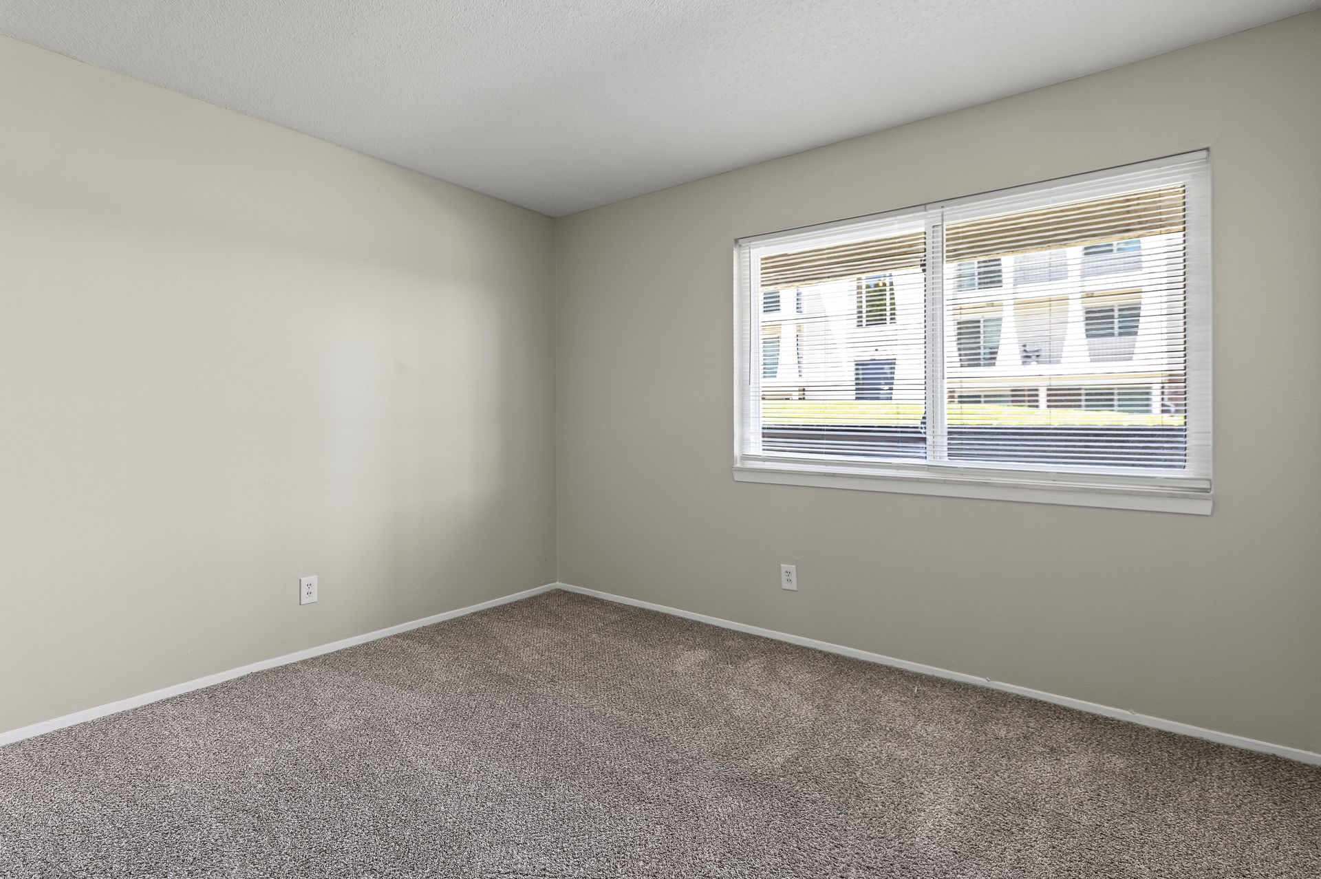 Empty room with beige walls, carpet, and a window looking out at a building.