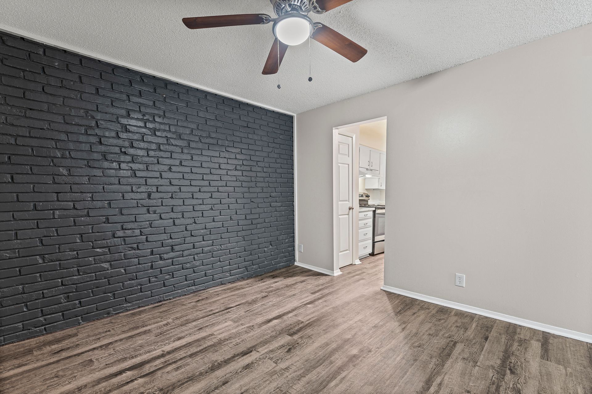 Empty room with painted black brick wall, wood floor, and doorway to a kitchen.