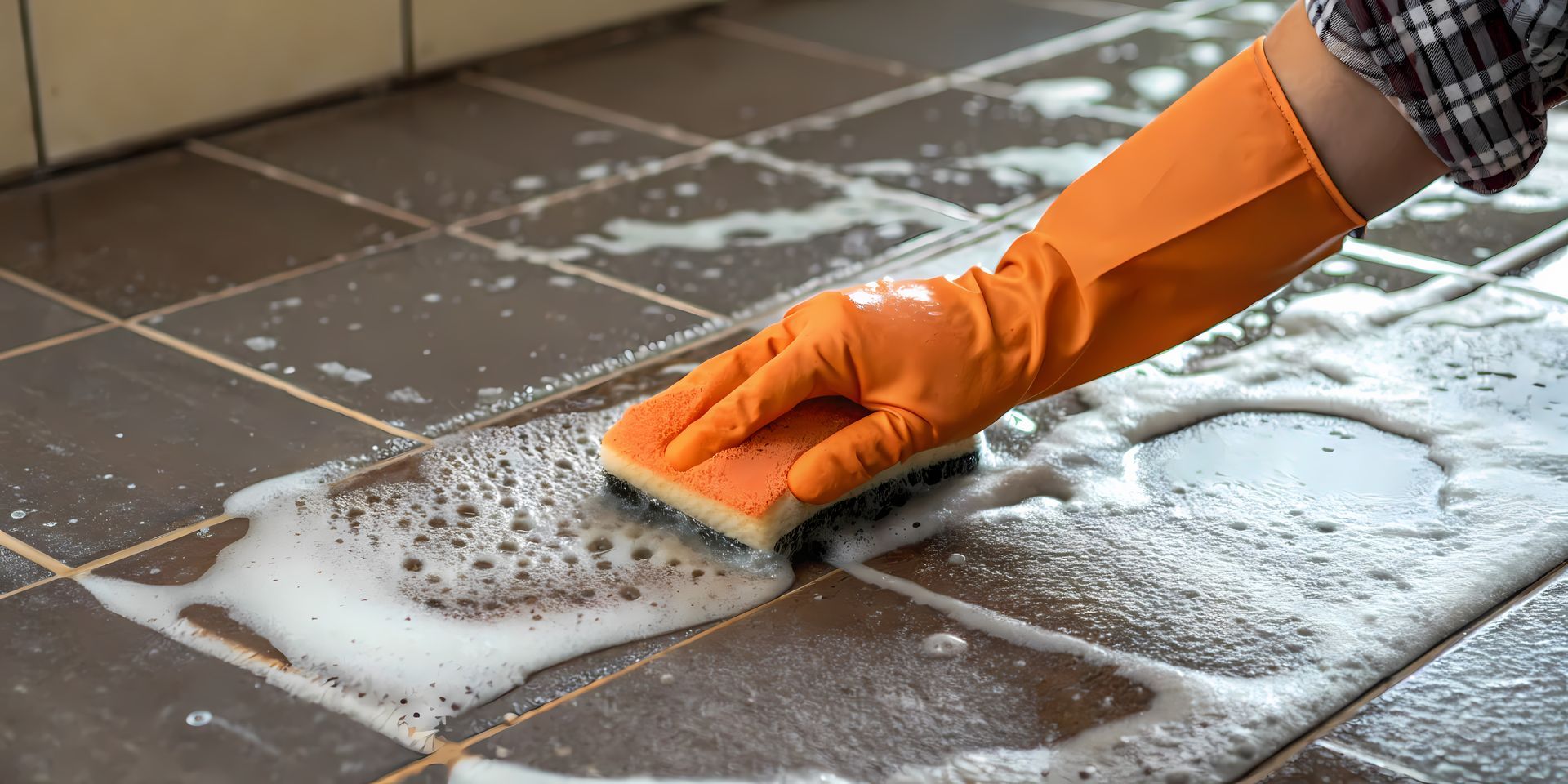 A person wearing orange gloves is cleaning a tile floor with a sponge.
