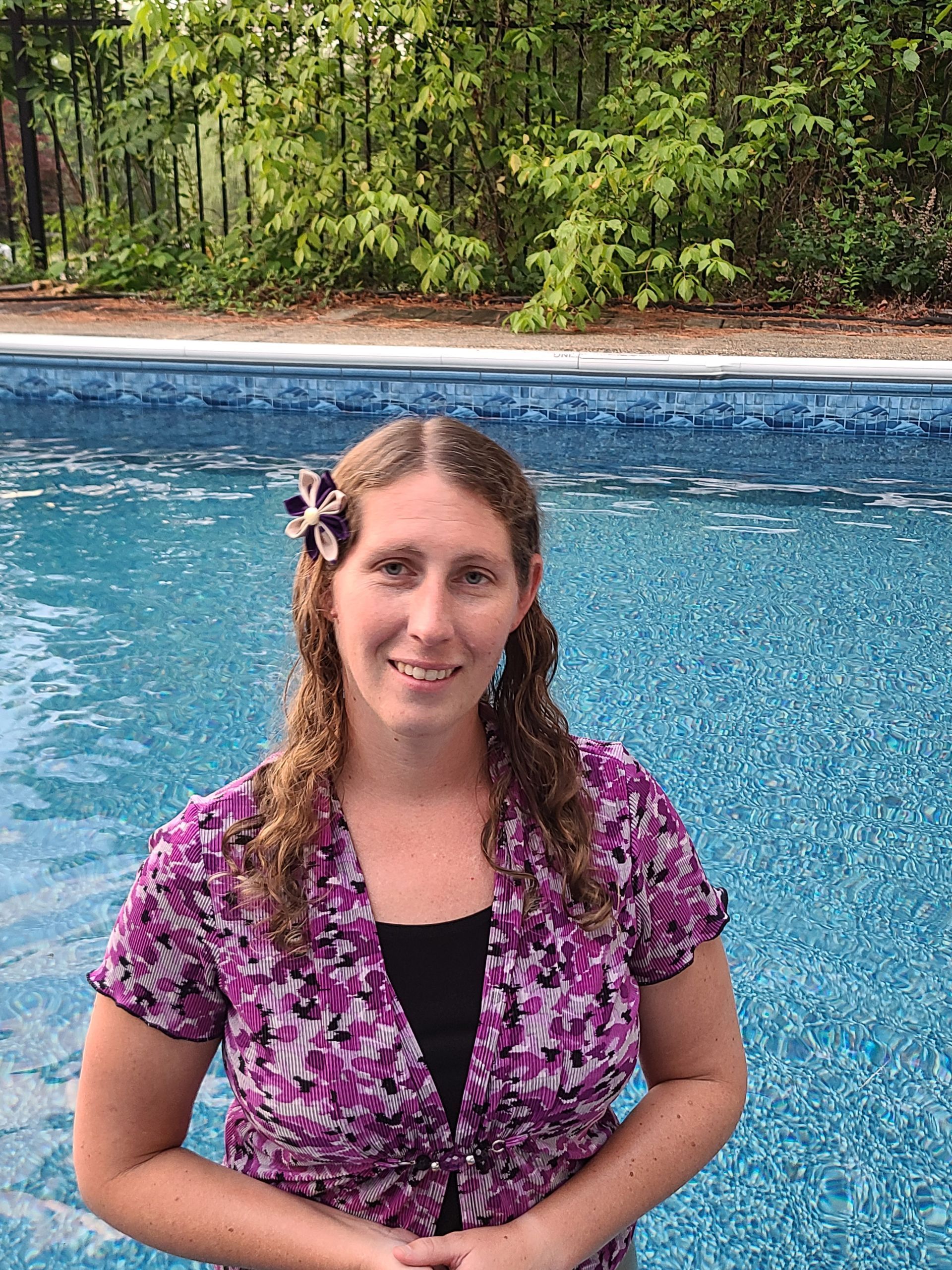 A woman in a purple shirt is standing in front of a swimming pool.