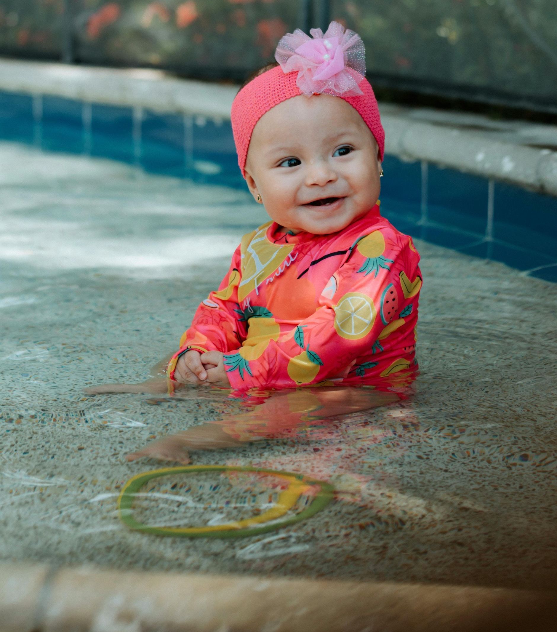 A baby is sitting in a swimming pool wearing a pink hat.