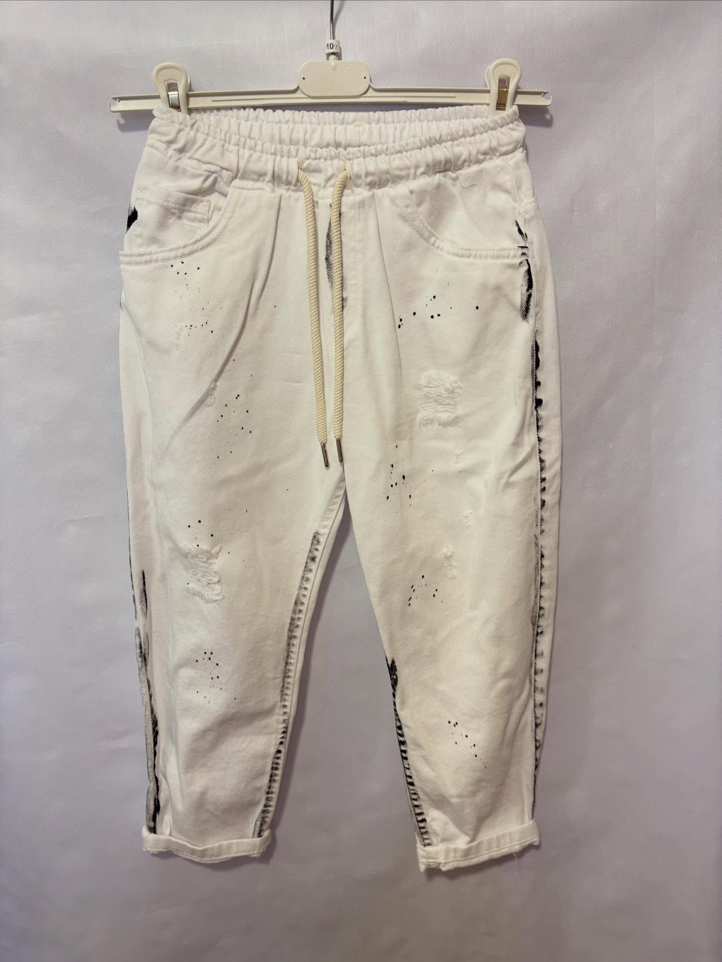 Pantaloni bianchi in denim corti con elastico in vita, coulisse e dettagli a schizzi di vernice, appesi a una gruccia.