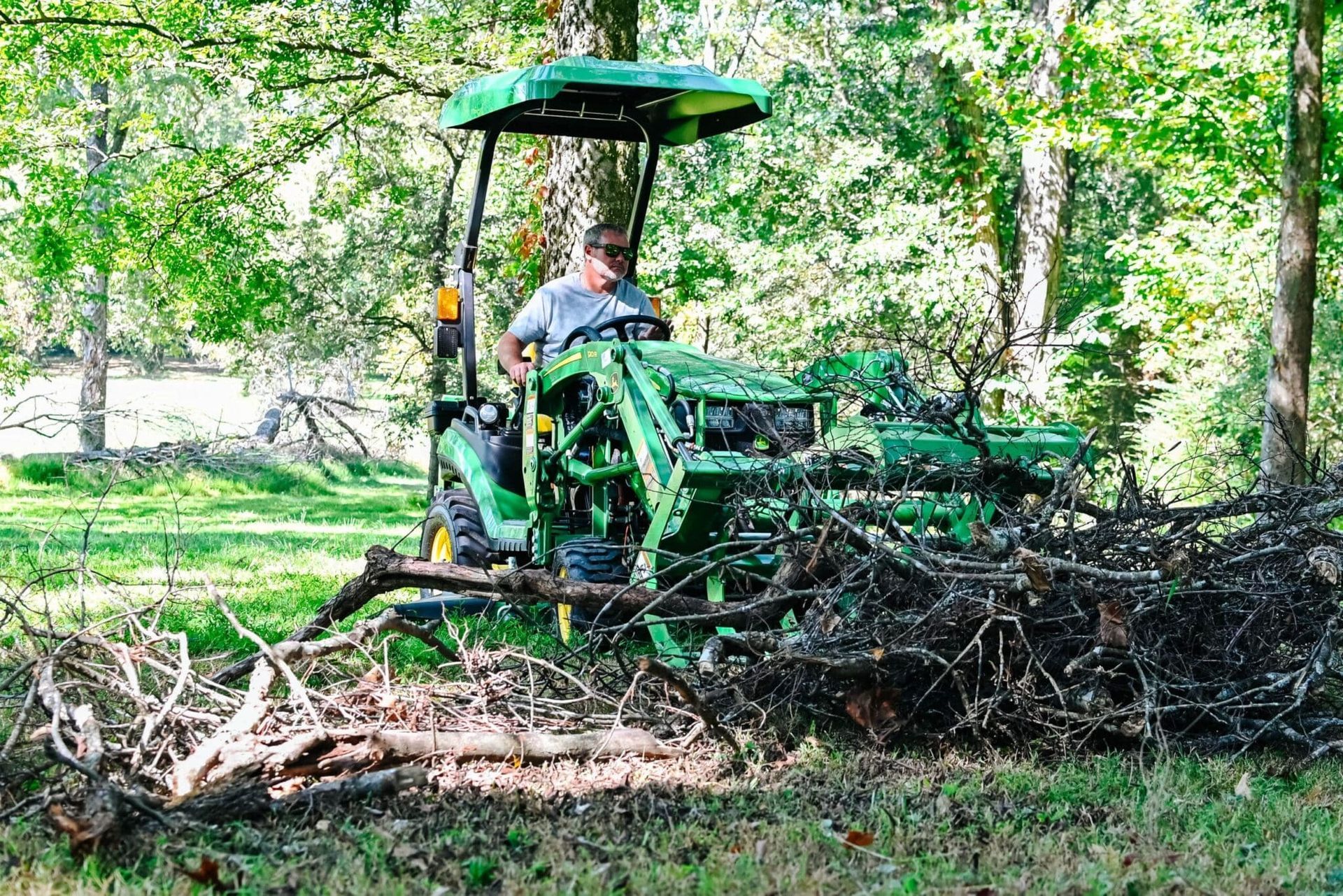 Man operating a green tractor, moving brush in a wooded area.