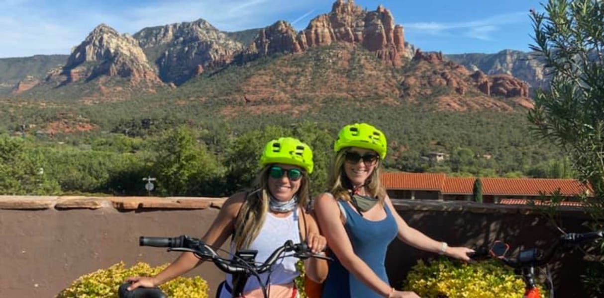 Two people wearing bright neon yellow bike helmets stand with bicycles against a backdrop of scenic red rock mountains.