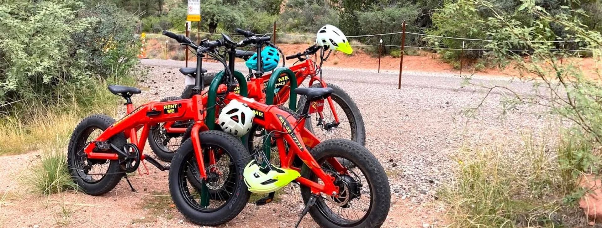 Three red fat-tire bikes with helmets parked on a gravel path beside vegetation. The setting appears to be an outdoor trail.