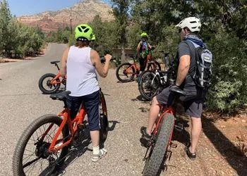 Group of people on e-bikes in a desert setting, wearing helmets. Red and orange bikes on a road.