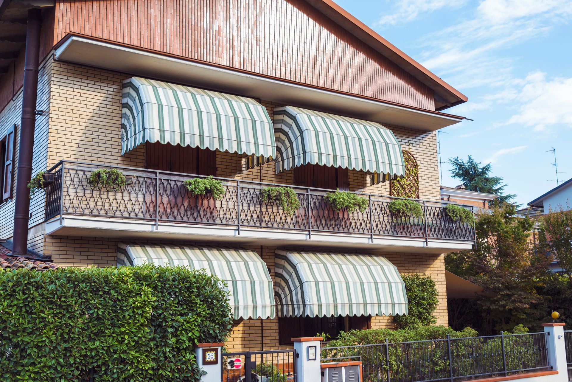 Two-story house with striped green awnings, brick exterior, balcony, and lush hedge in front