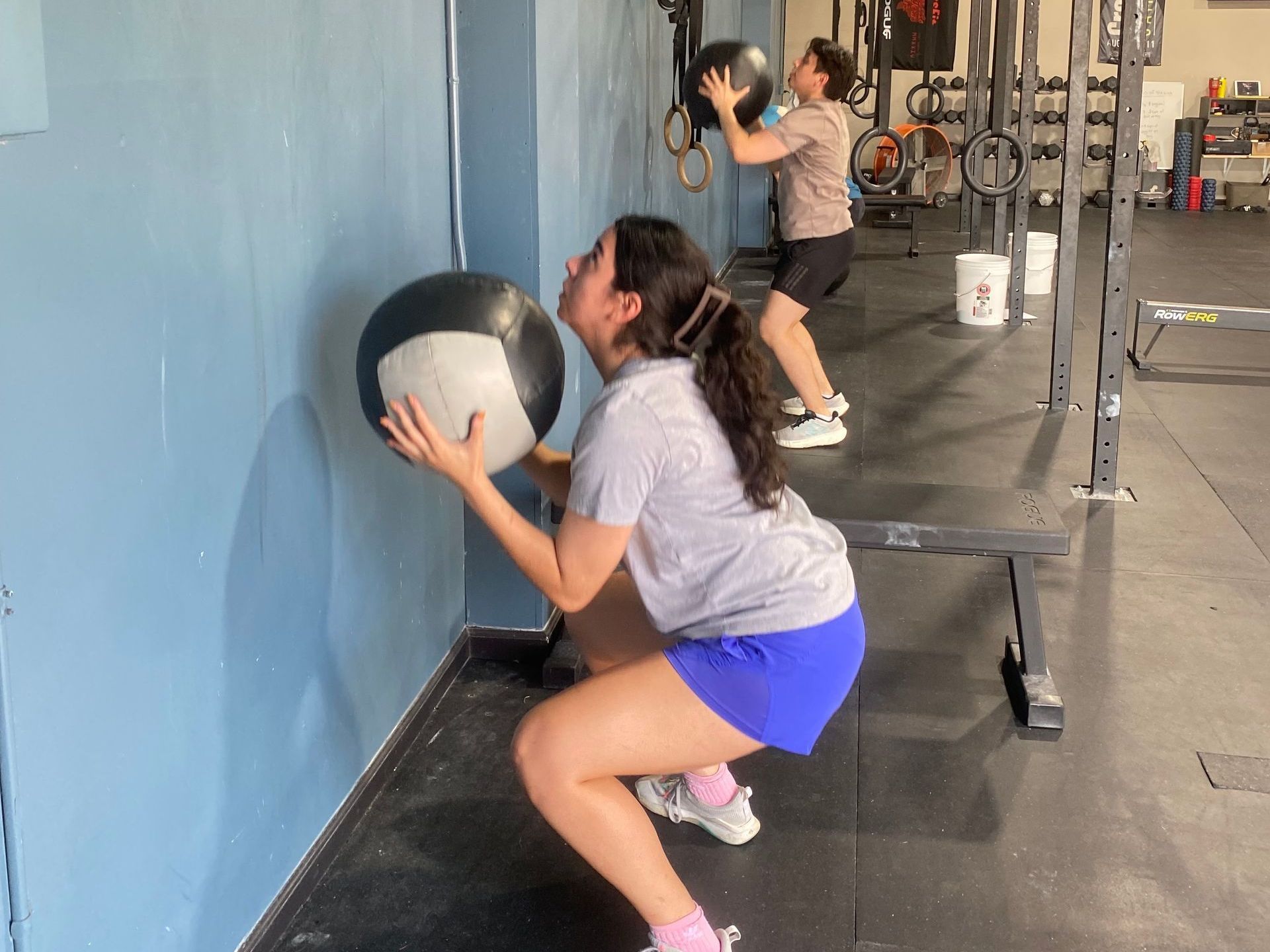 A woman is squatting down while holding a medicine ball in a gym.