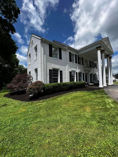 White two-story house with black shutters, large columns on a porch, and lush green lawn under a partly cloudy sky.