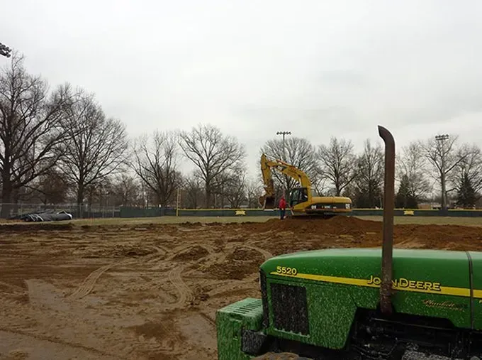 A green john deere tractor is parked in a dirt field with a yellow excavator in the background.