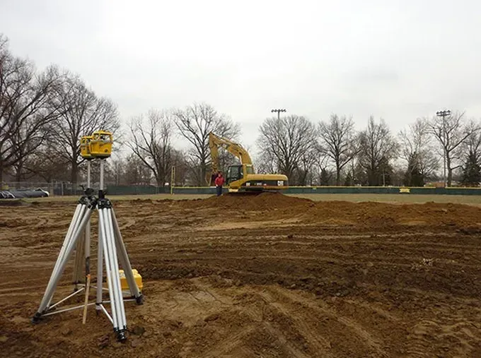 A construction site with a yellow excavator in the background and a tripod in the foreground.