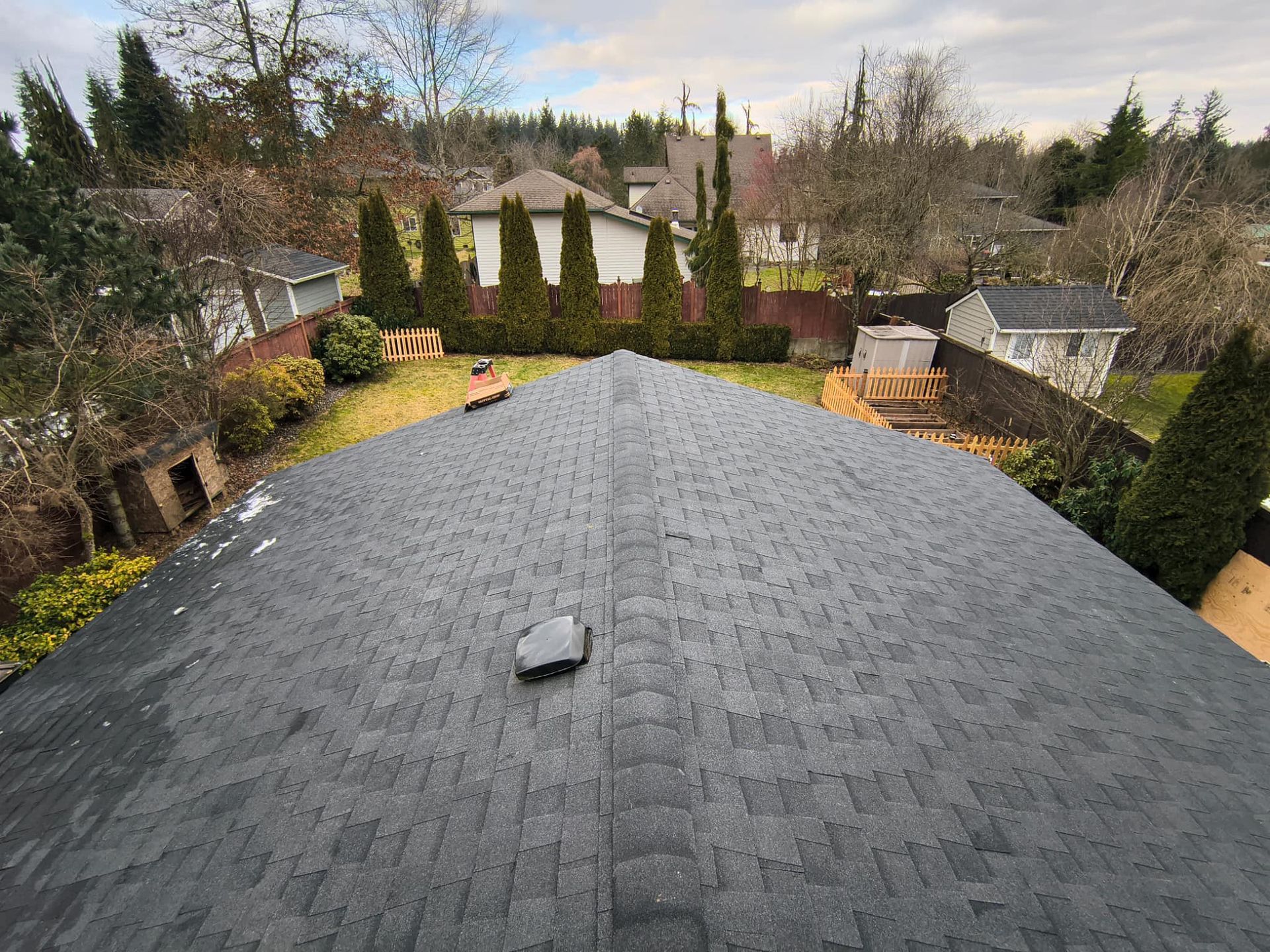 View of a dark shingle roof with a vent, overlooking a backyard with houses and trees on a cloudy day.
