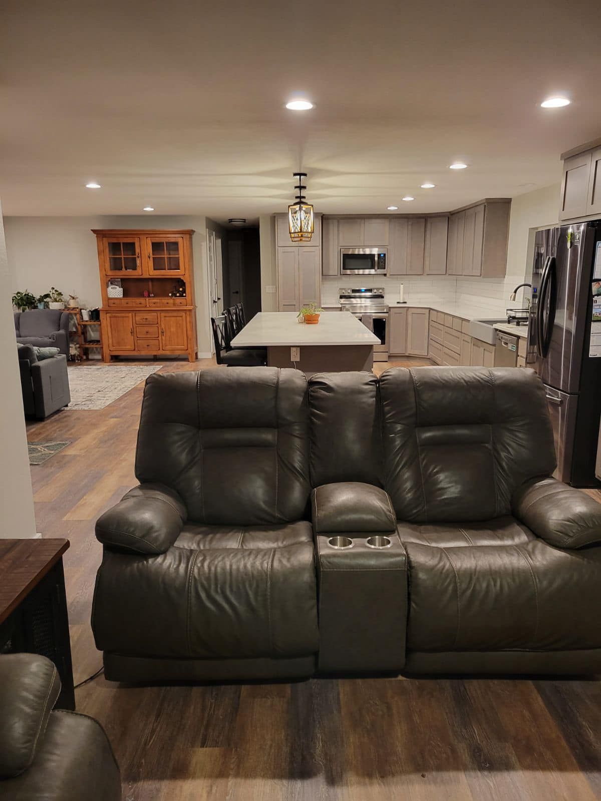 Living room with dark gray reclining sofa, leading to open kitchen with island and light wood cabinets.