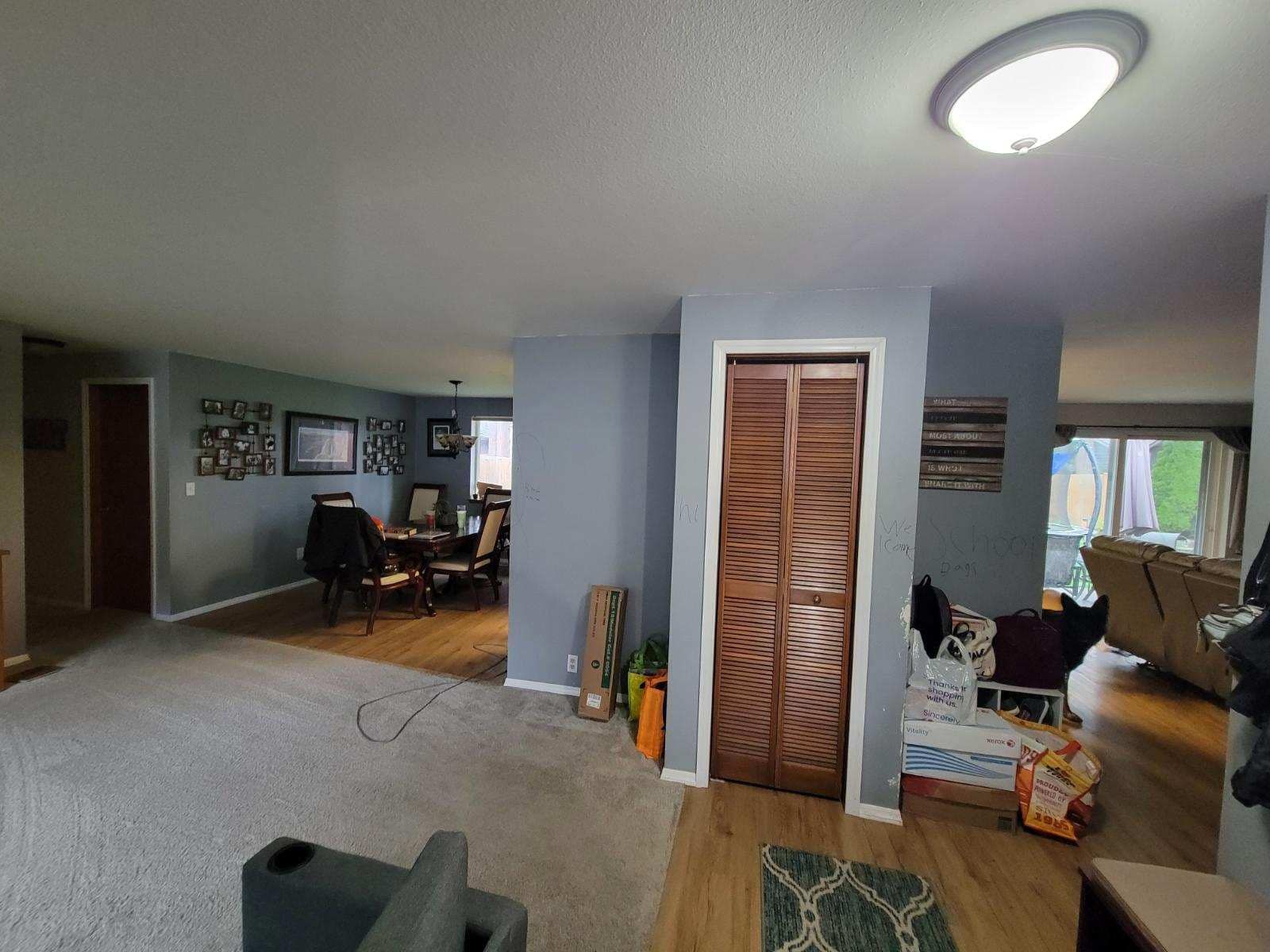 A view of a living room with gray carpet, blue walls, a dark wooden closet, and a dining room in the background.