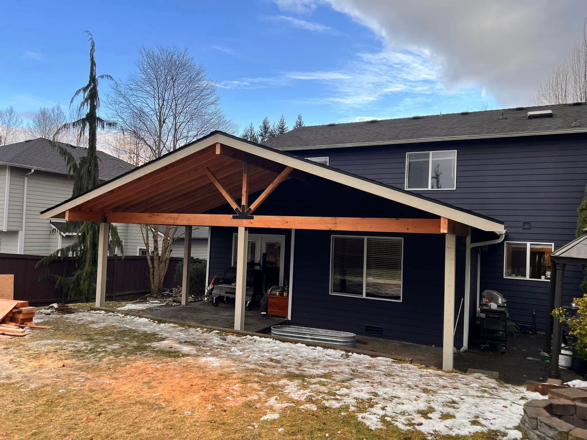 Backyard with a dark blue house and a newly built wooden patio cover. Snowy ground, sunny sky.