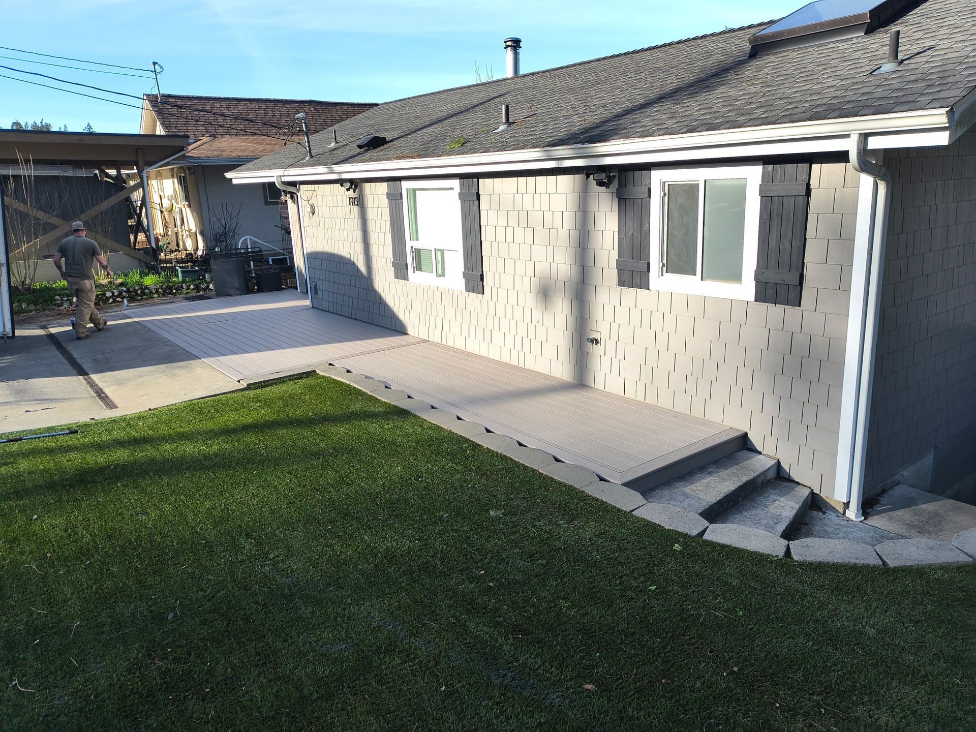 A light gray house with a small deck and steps leading to a green lawn. A person is near the driveway.