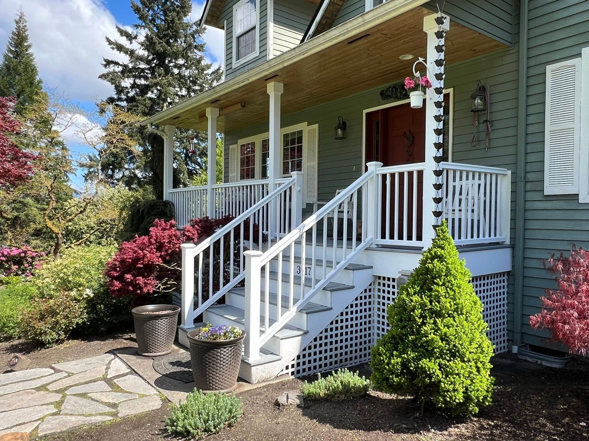 Green house with white porch, stairs, and railing, surrounded by bushes and a stone path.