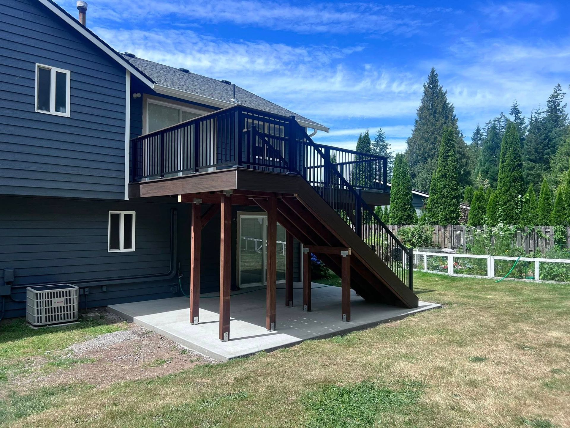 Two-story home with dark blue siding, a deck, and stairs leading to a concrete patio in a grassy yard.
