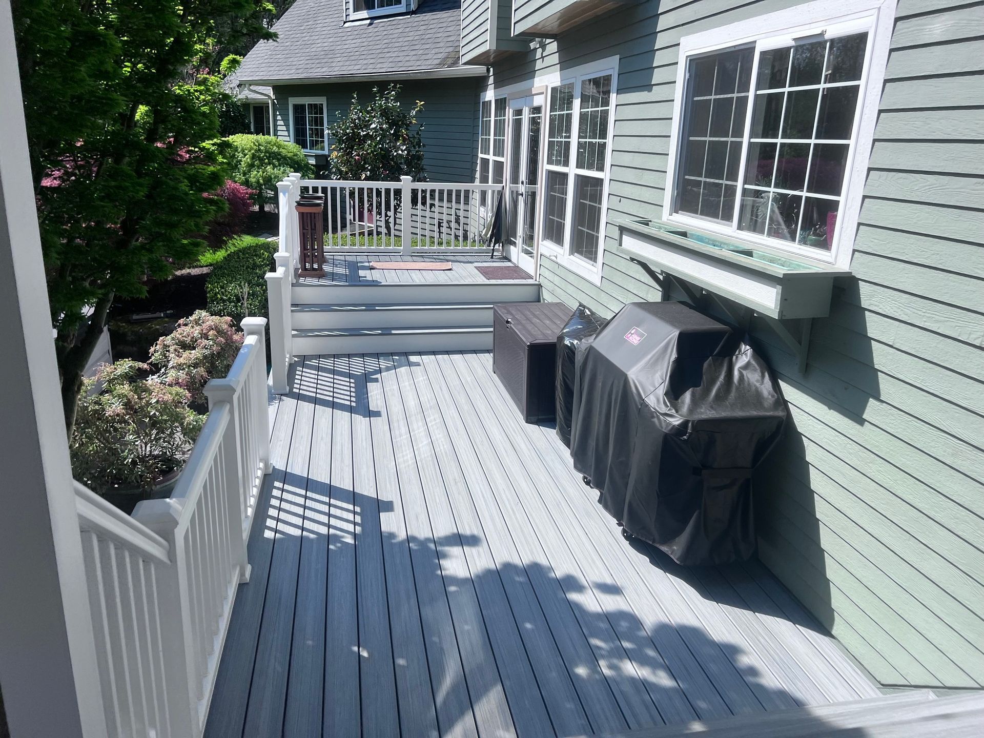 Deck with grill and white railings next to a green house, sunny day.