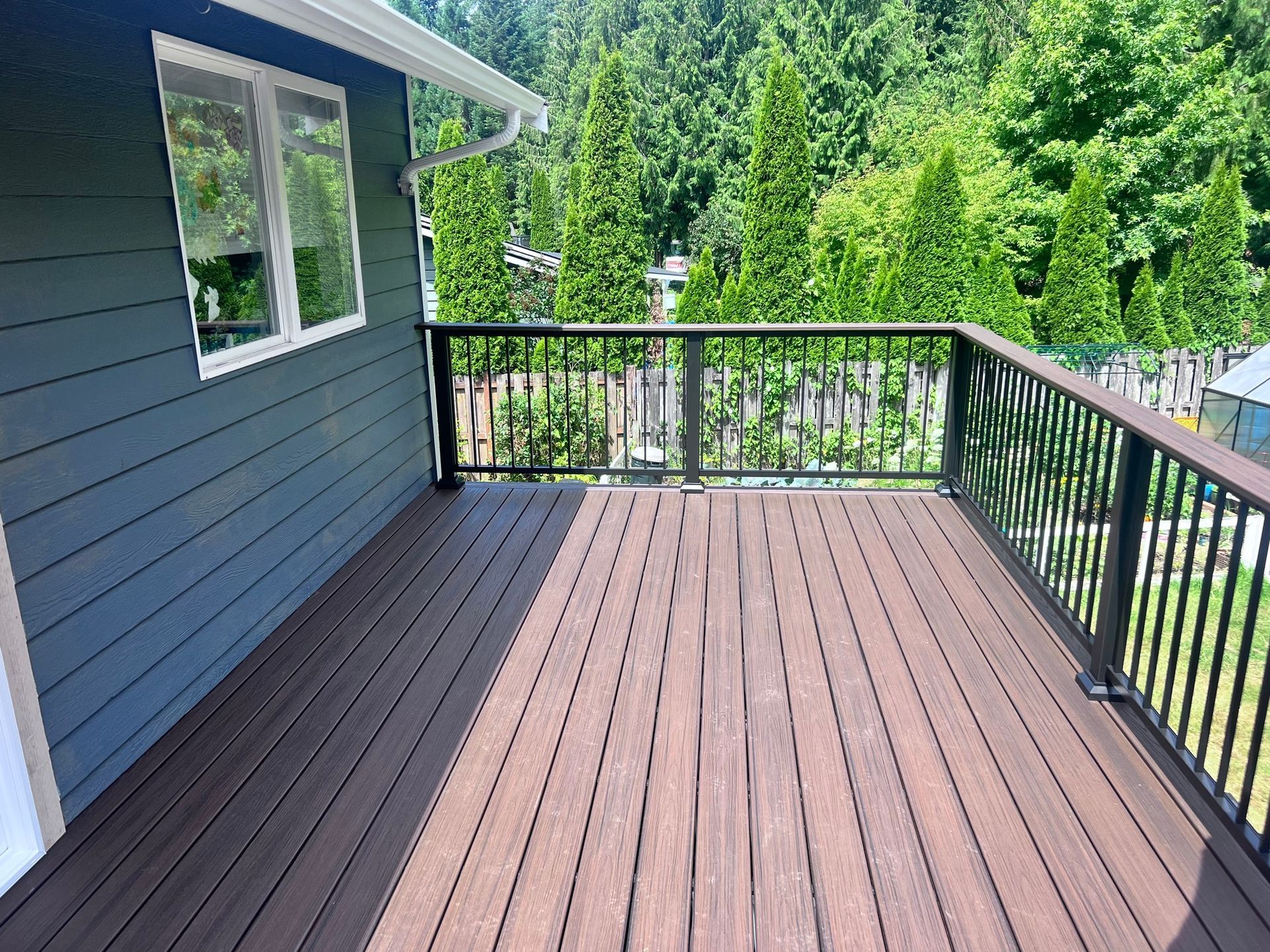 Brown composite deck with black railing and blue-gray siding, overlooking a wooded backyard.