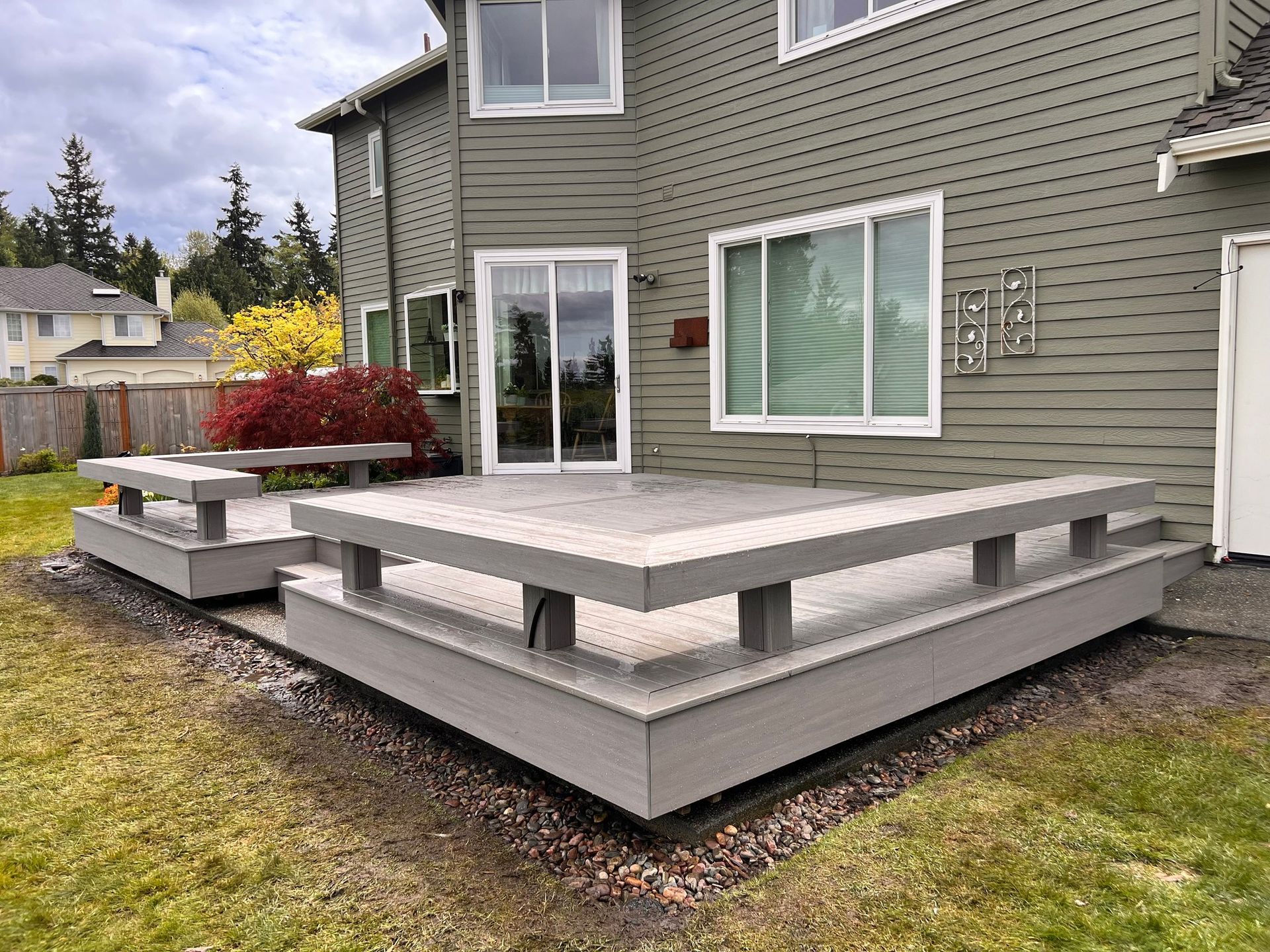 Gray composite deck attached to a house with a sliding glass door and windows. Lawn in foreground, trees in background.