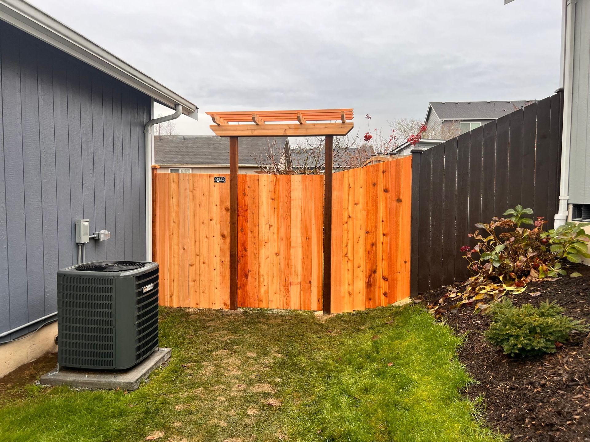 Wooden fence with gate, enclosing yard between two houses. AC unit on the left. Green grass.