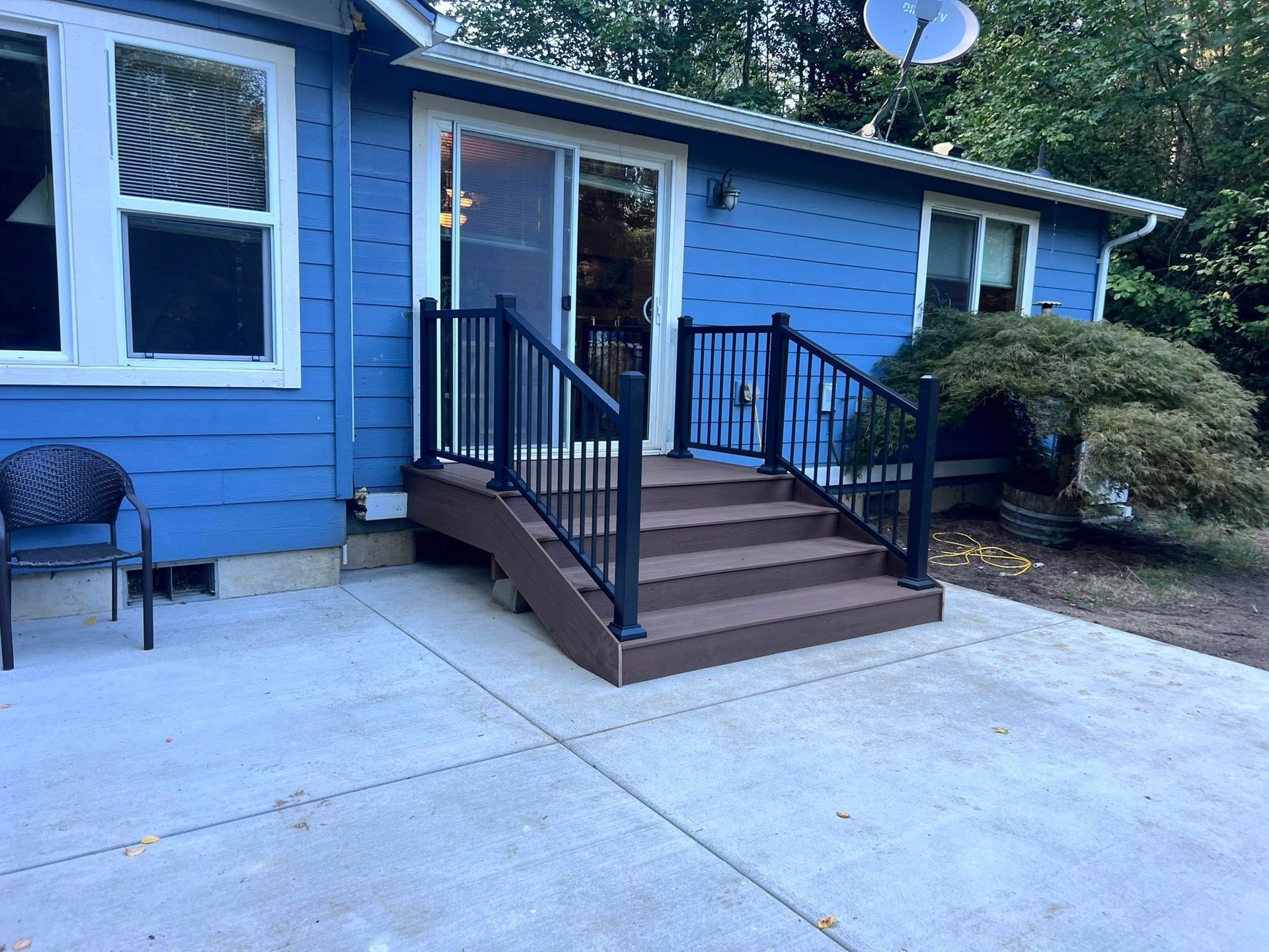 Blue house with concrete patio, steps up to a deck with black railing.