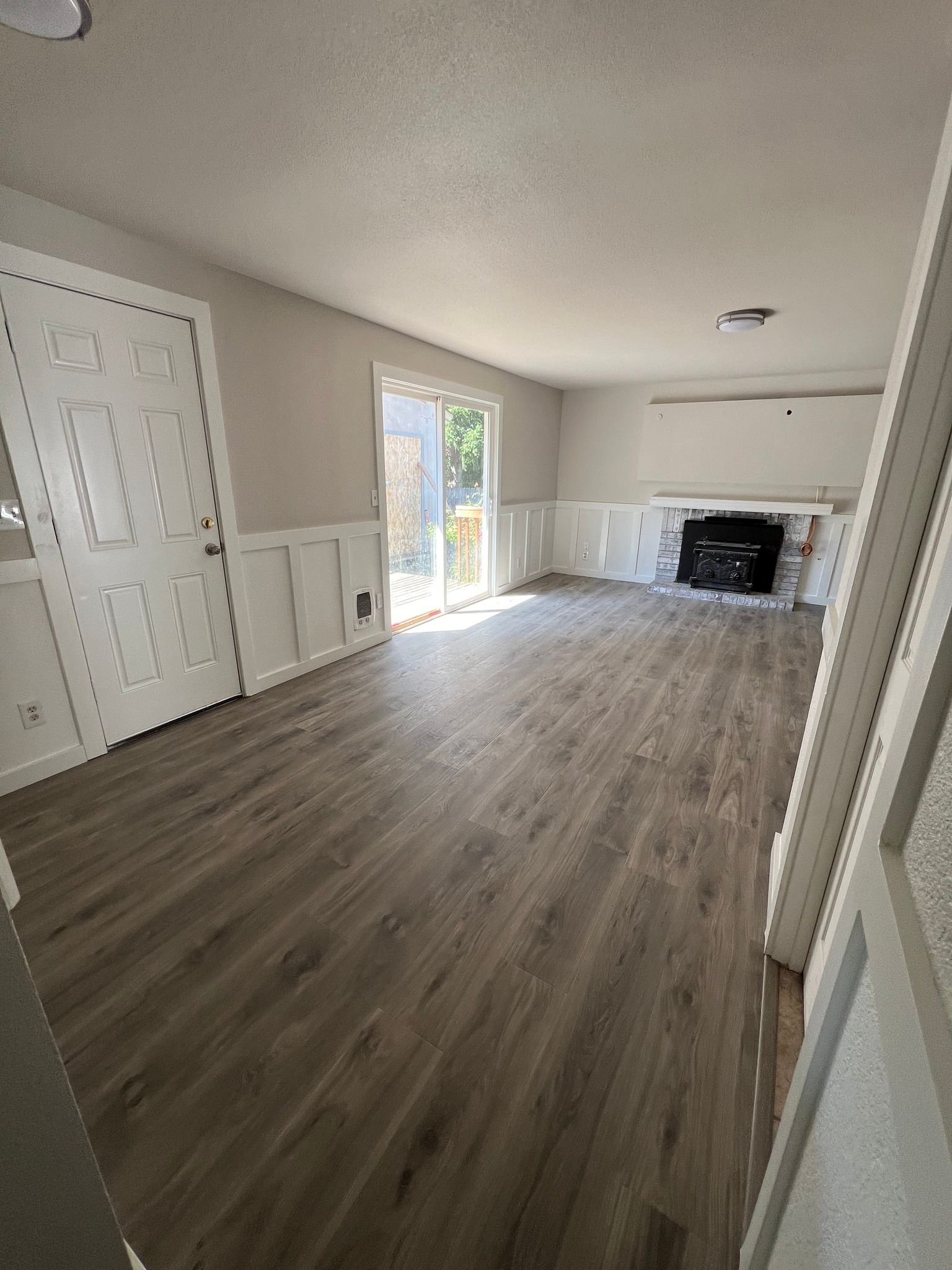 Empty living room with wood-look flooring, white walls with wainscoting, sliding door, fireplace, and closed white door.