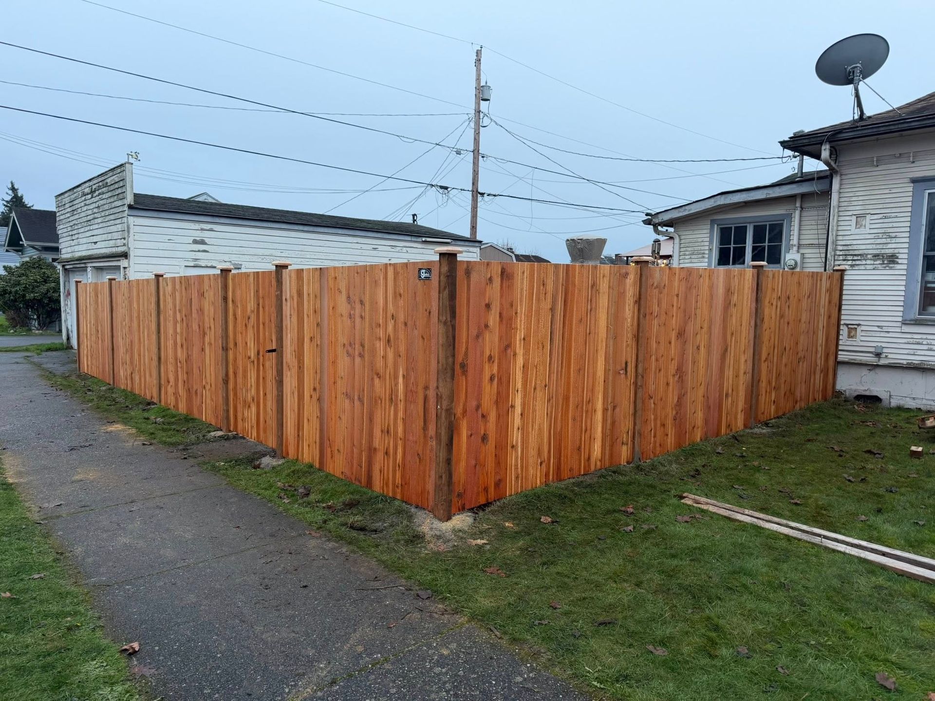 A newly built wooden fence surrounds a green lawn next to a paved alley and small houses under a cloudy sky.