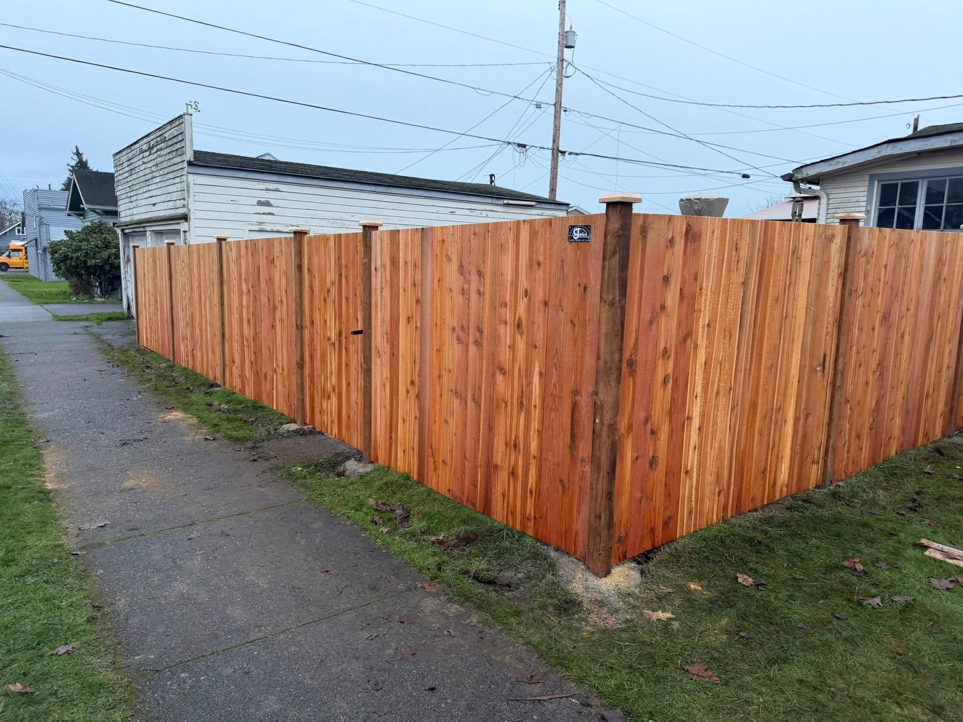 Wooden fence corner along a sidewalk, grass, and houses; overcast day.