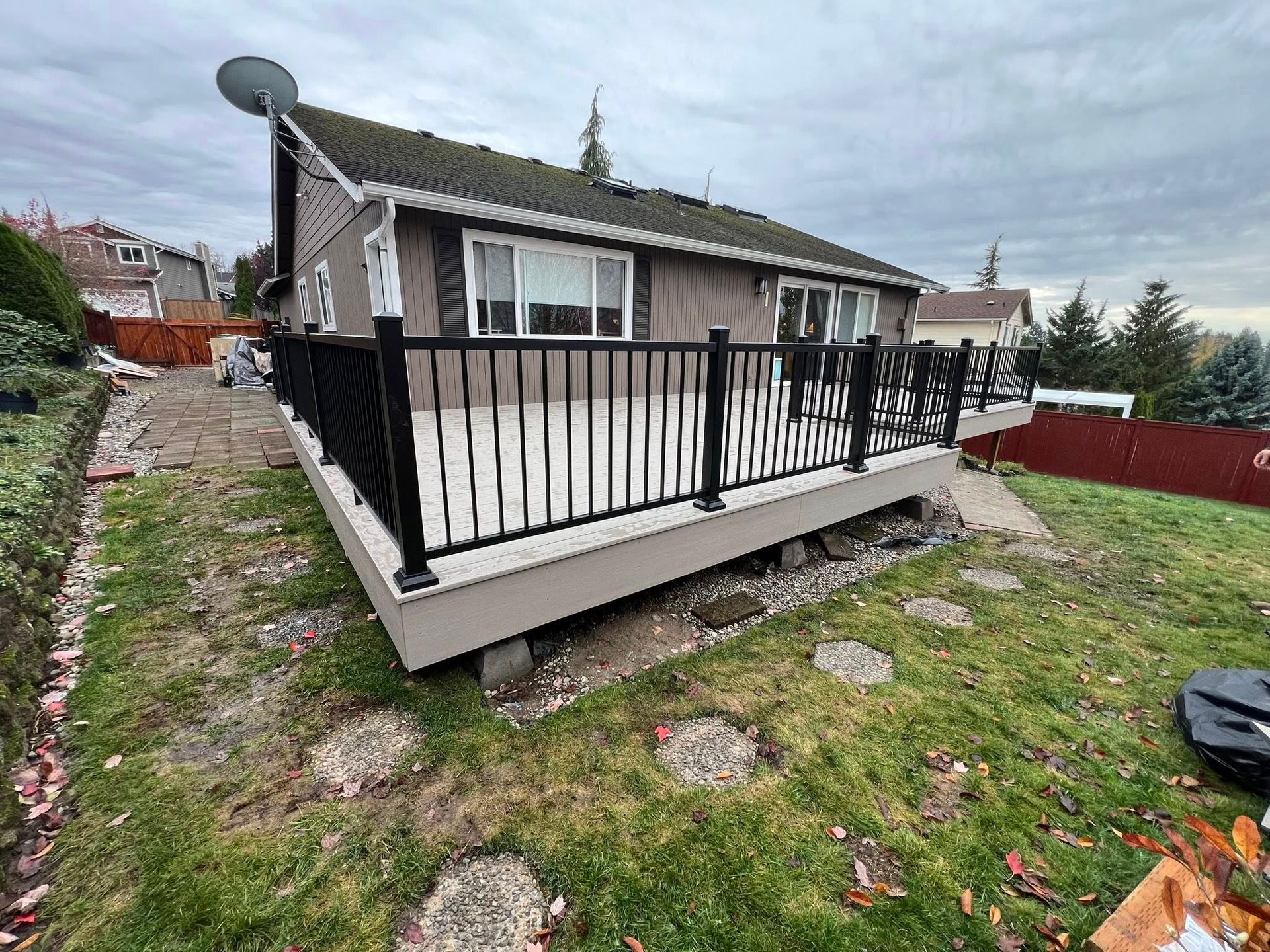 A house with a black railing deck overlooking a grassy yard with stepping stones under an overcast sky.