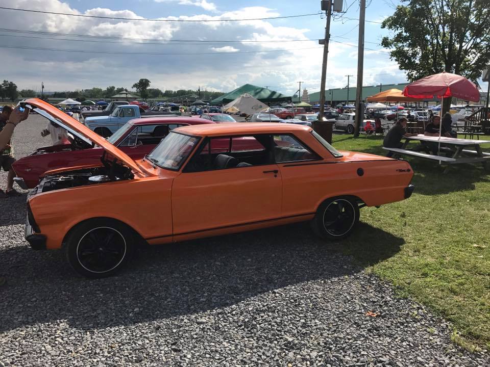 An orange car with the hood up is parked in a gravel lot