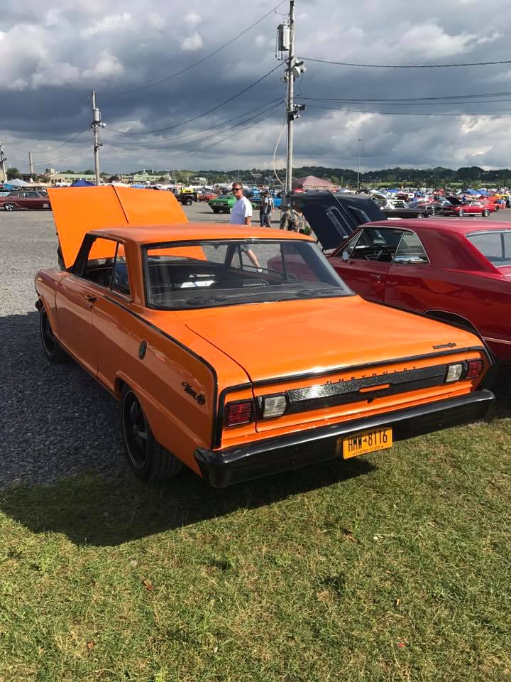 An orange car with the hood up is parked in a grassy field.
