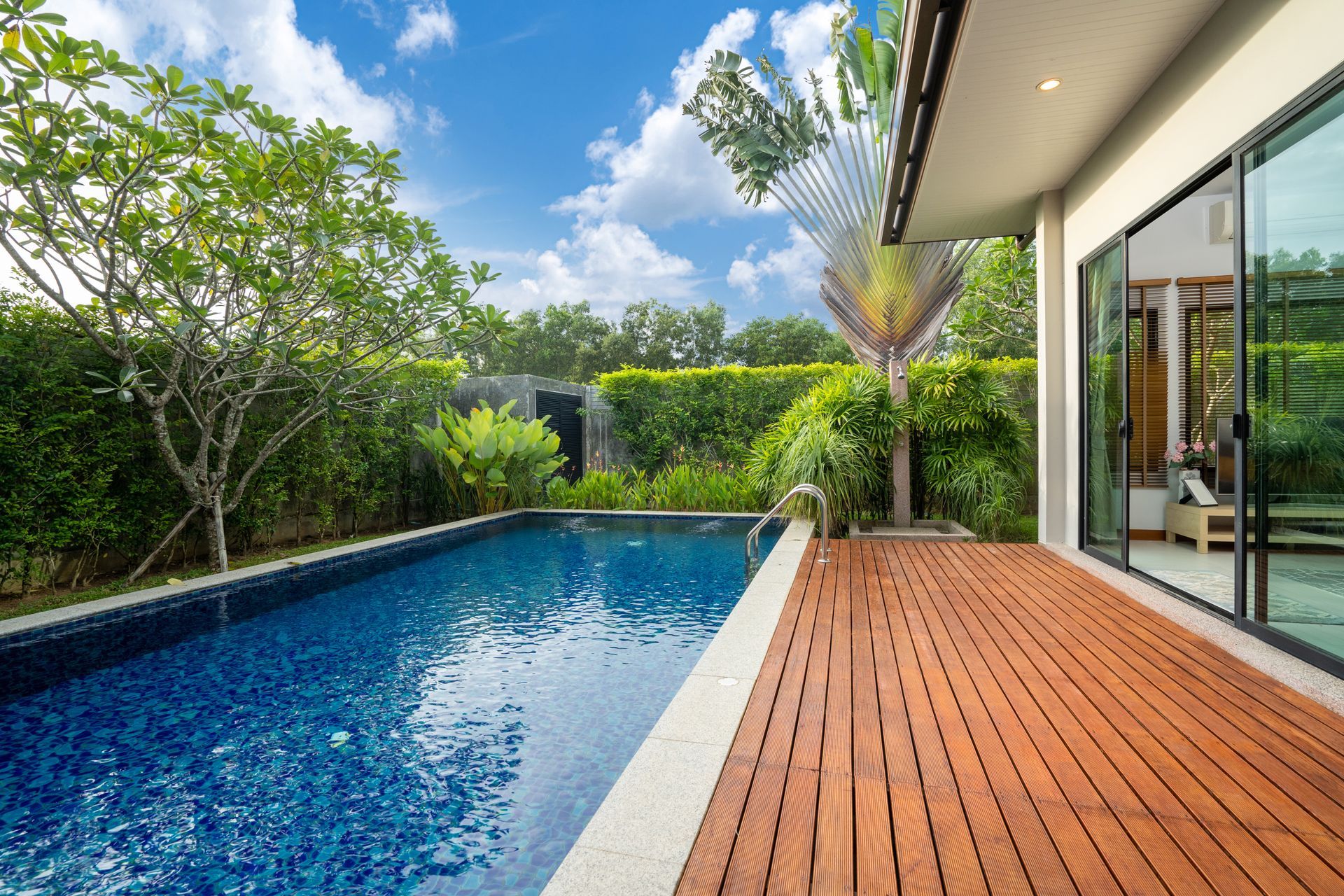 Modern backyard pool with blue water, wooden deck, and lush greenery beside a house.