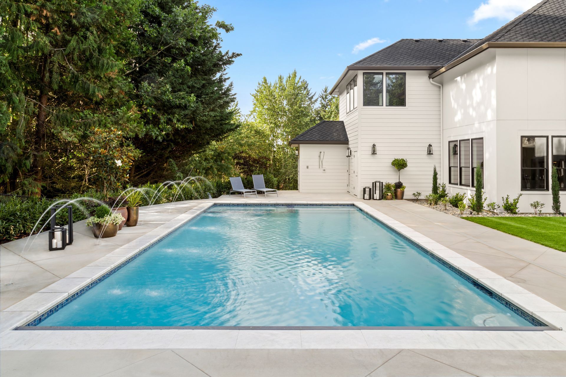 Rectangular backyard swimming pool with fountain jets beside a modern two-story home.