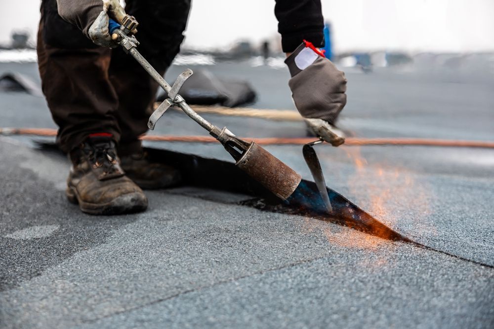 Roofer using a torch to seal a seam on a dark, flat roof. Flames are visible.