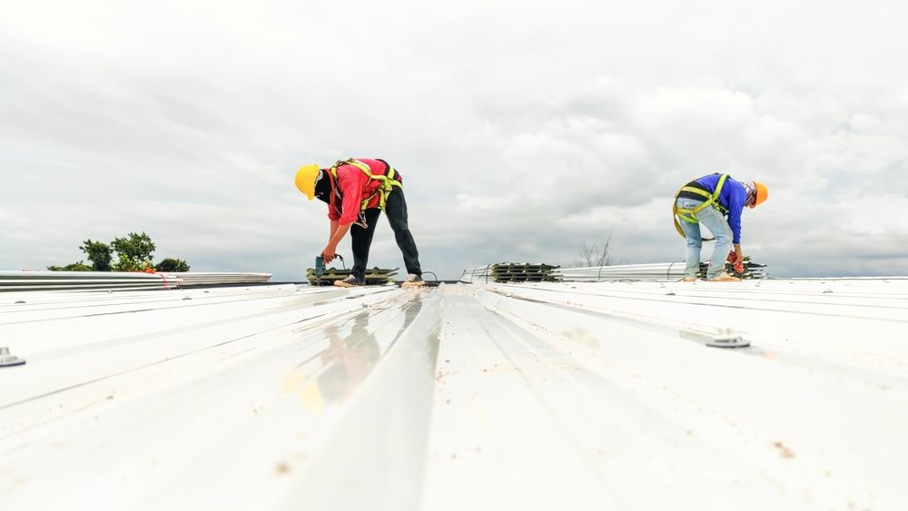 Two roofers in harnesses on a white metal roof, working under a cloudy sky.